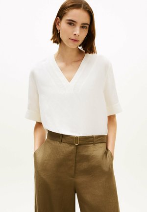 Frau mit kurzen braunen Haaren, die ein weißes V-Ausschnitt-Kurzarmshirt trägt, das in hoch taillierte, olivgrüne Hosen mit Schnallengürtel gesteckt ist.