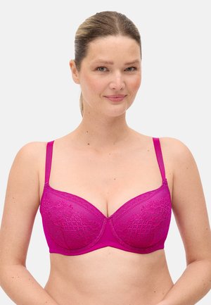 Mujer de piel clara y cabello rubio sonríe, lleva un sujetador de encaje rosa brillante con tirantes ajustables delgados sobre un fondo blanco liso.