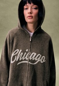 Žena s krátkymi čiernymi vlasmi oblečená v hnedej mikine na zips s nápisom "Chicago" na prednej strane, stojaca pred jednofarebným pozadím.