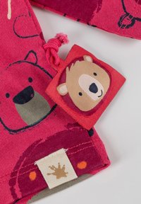 Rosa Stoff mit Bärenillustrationen, einem gewebten Etikett mit einem Bärenkopf und einem beigen Label mit einem braunen Spritzgrafik. Strukturierter und farbenfroher Entwurf.