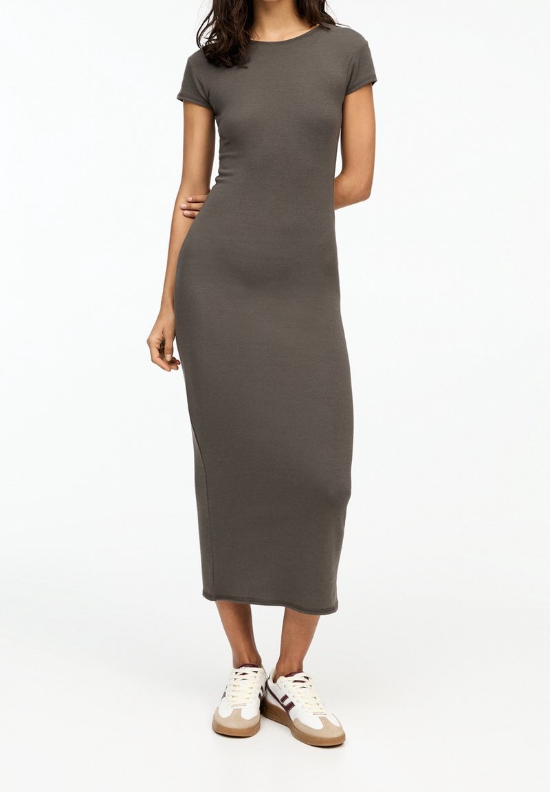 Femme portant une robe midi marron ajustée à manches courtes et des baskets blanches, debout avec une main sur la hanche sur un fond uni.