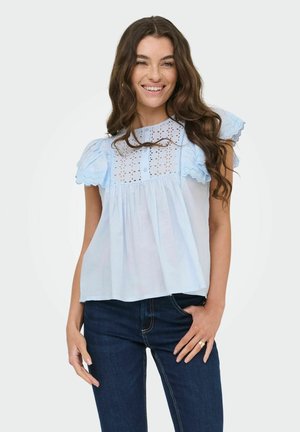 Femme aux longs cheveux bruns souriant, portant un chemisier bleu clair à broderie anglaise et manches à volants et un jean bleu foncé, debout devant un fond uni.