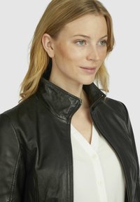 Black leather jacket with zip closure, high collar, and smooth texture, worn over a white button-up shirt. Features minimal accents.