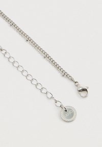 Srebrna metalowa naszyjniczka z ogniwami i siateczkowanymi sekcjami. Posiada zapięcie typu lobster oraz okrągłą zawieszkę z wyrytym logo marki.