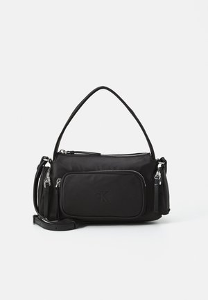 Sac à bandoulière noir avec poignée courte et sangle réglable, poche zippée devant et logo embossé "cK" sur fond blanc uni.