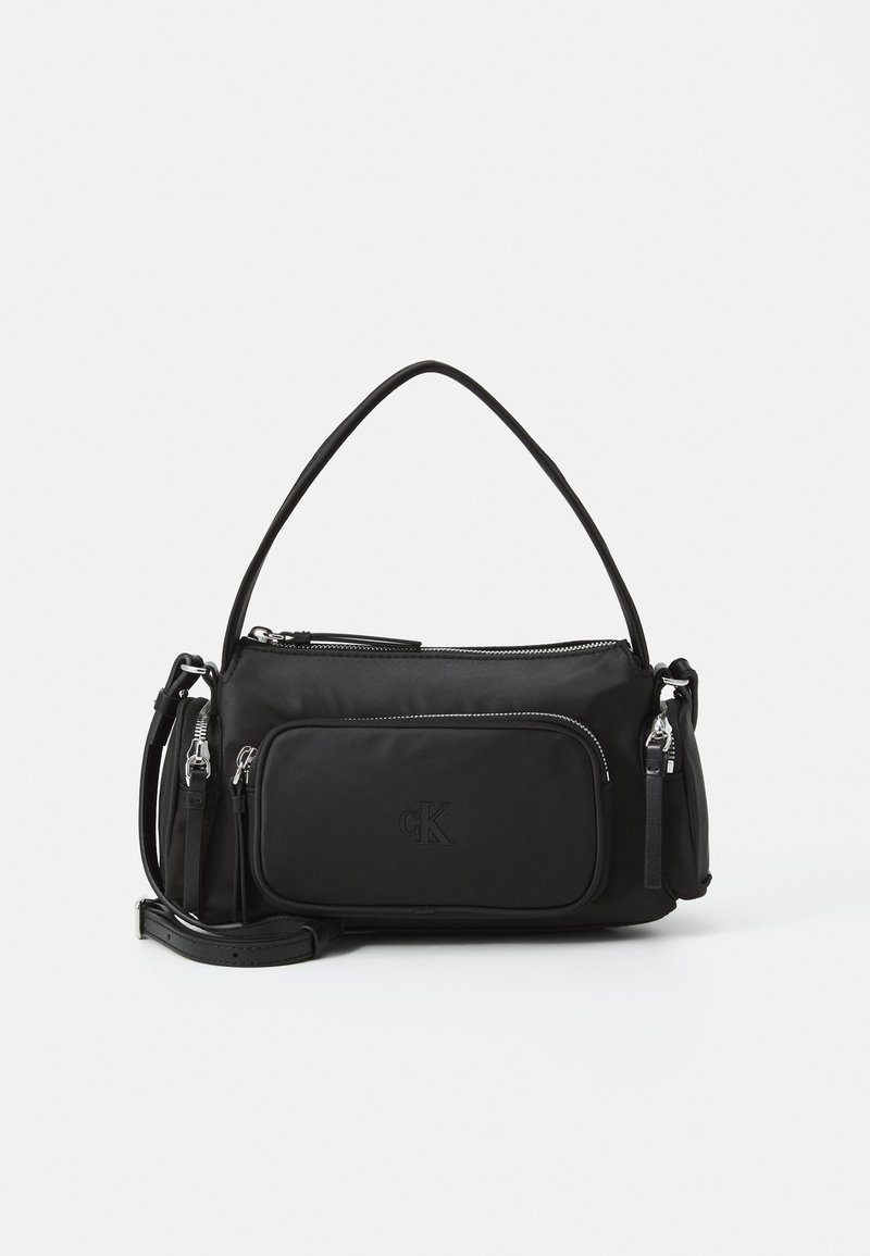 Sac à bandoulière noir avec poignée courte et sangle réglable, poche zippée devant et logo embossé "cK" sur fond blanc uni.