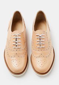 Beige nahkakengät brogue-tyyliin, joissa on rei'itettyä yksityiskohtaa, pyöreä kärki ja valkoiset nauhat. Ominaisuuksina teksturoidut pinnat ja kontrastivärinen ruskea pohja.