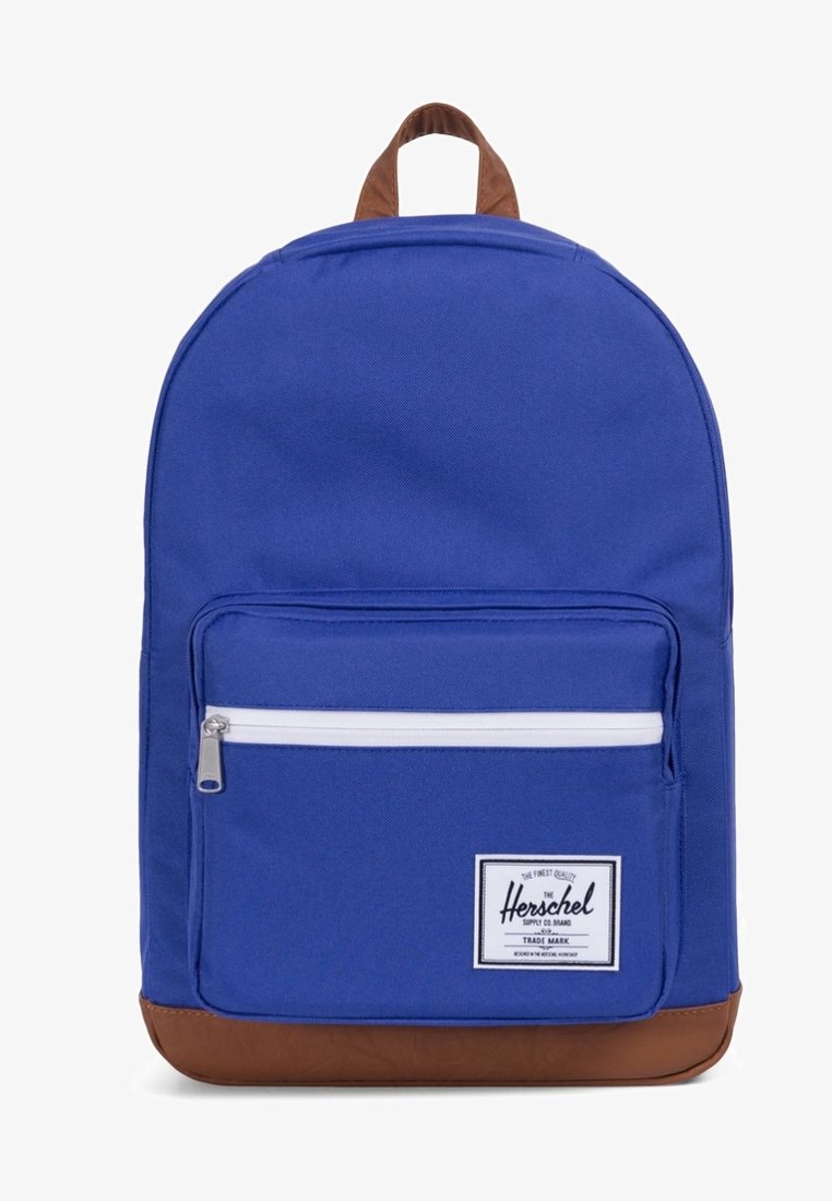 Sac à dos en tissu bleu avec une base et une poignée en synthétique marron, doté d'une poche avant zippée et d'une fermeture éclair blanche. Patch logo à l'avant.