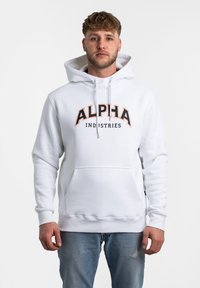 Sudadera blanca con un bolsillo frontal, capucha con cordón y el texto audaz "ALPHA" en negro y naranja. Textura suave, diseño casual, combinada con jeans de color azul claro.