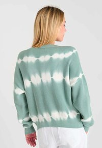 Groene geribbelde gebreide trui met wit tie-dye patroon, losse pasvorm, verlaagde schouders en lange mouwen. Witte broek zichtbaar daaronder.