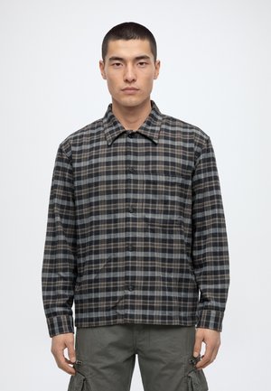 Un homme se tient debout, face à l’avant, devant un fond uni. Il porte une chemise boutonnée à carreaux foncés et un pantalon cargo gris.