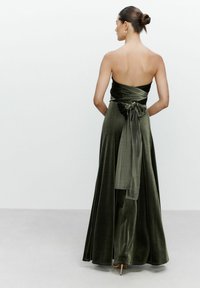 Eine Frau mit einem Haarknoten trägt ein trägerloses, dunkelgrünes Samtkleid mit einer großen Schleife, die um die Taille gebunden ist, während sie vor einem schlichten Hintergrund steht.
