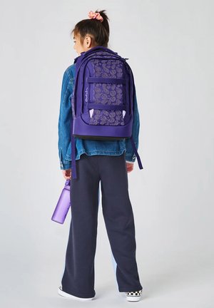 Child wearing a denim jacket and dark pants, carrying a purple backpack with patterns and holding a matching purple water bottle.