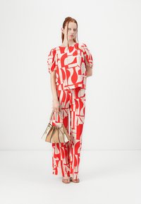 Ensemble à motifs rouge et blanc comprenant un top à manches bouffantes et un pantalon large. Le mannequin tient un sac à main beige au design froncé.