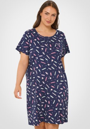 Kvinde iført en navyblå kortærmet kjole med farverige slik- og slikkepindemønstre og en knappet halsudskæring, stående mod en hvid baggrund.