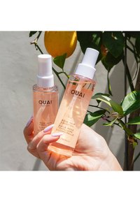 To flasker med OUAI hår- og kroppsolje i klare flasker med hvite pumper. Begge inneholder en rosafarget væske, merket med svart tekst. Hånd som holder flaskene.