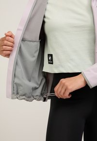 Leichte Jacke mit grauem Innenfutter, rosa Akzenten, verstellbarem Kordelzugsaum und einer Tasche. Getragen über einem hellgrünen Oberteil. Schwarzes Logo-Tag sichtbar.