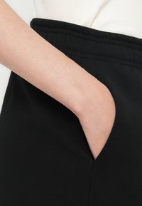 Hånden plassert inne i lommen med svart elastisk midjebånd på casual bukser, brukt med hvit ribbet topp.