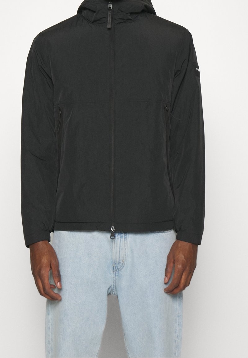 Svart, lätt zip-up jacka med huva, som har två sidfickor och en mjuk textur. Bärs över ljusblå denimbyxor.