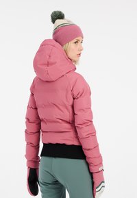 Rosa Pufferjacke mit Kapuze und geripptem schwarzen Saum. Trägt dazu grüne Hosen und gestreifte, gemusterte Handschuhe. Weiche Textur und strukturiertes Design.