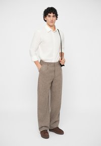 Ung mann med krøllete hår iført en hvit langermet skjorte, bredbente bukser i tweed, brune joggesko og en crossover-veske, som står mot en hvit bakgrunn.