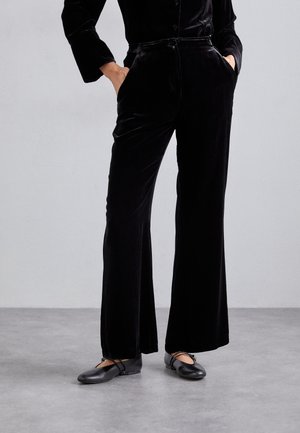 Persona con pantalones de terciopelo negros de talle alto y pierna ancha y zapatos de ballet negros, de pie con las manos en los bolsillos sobre suelo gris.