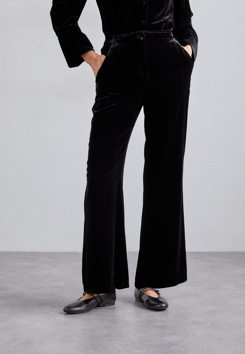 Personne portant un pantalon en velours noir taille haute à jambes larges et des ballerines noires, debout les mains dans les poches sur un sol gris.