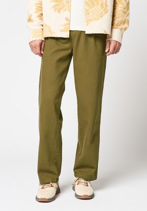 Homme portant un pantalon droit kaki, un cardigan en maille crème et beige avec un motif floral, et des baskets crème avec des semelles vertes.