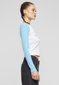 Biely crop top s dlhými svetlomodrými rukávmi, s jemne vrúbkovanou textúrou a potlačeným nápisom vpredu. Skombinovaný s tmavými džínami.