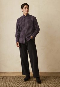Lilla plaid skjorte med knaplukning og krave, parret med brede sorte bukser. båret med mørke sko, sat mod en neutral baggrund.