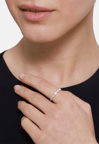 Srebrny pierścionek z serią okrągłych, polerowanych akcentów. Noszony na palcu, kontrastując z czarną bluzką. Gładka faktura i minimalistyczny design.