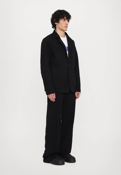 Blazer noir en tissu texturé avec col à revers, fermeture à un bouton, et deux poches avant, associé à un pantalon noir ample.