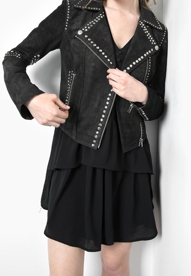 Femme ajustant une veste de biker noire cloutée sur une robe noire superposée, debout devant un fond blanc.
