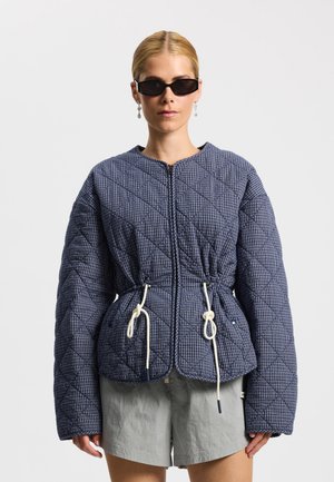 Kobieta w czarnych okularach przeciwsłonecznych, niebieskiej pikowanej kurtce w kształcie diamentu z białymi sznurkami oraz jasnoszarych szortach na białym tle.