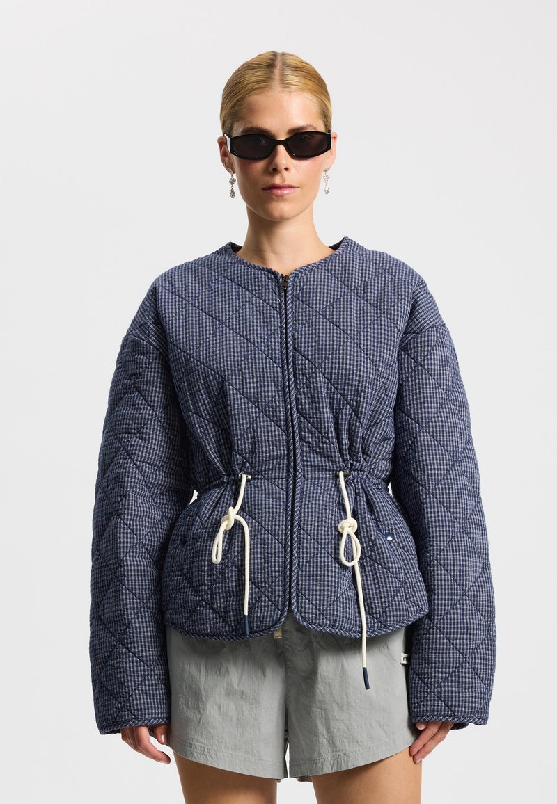 Žena s čiernymi slnečnými okuliarmi, modrou prešívanou bundou v tvare diamantu s bielymi sťahovacími šnúrkami a svetlosivými šortkami na bielom pozadí.