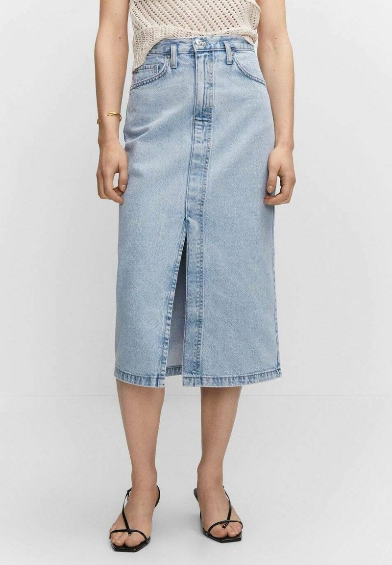 Femme portant une jupe midi en denim bleu clair avec une fente devant, associée à un top en tricot beige et des sandales noires à brides.