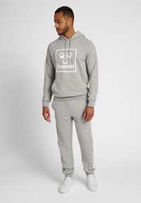 Grijze hoodie met een wit logo, bijpassende grijze sweatpants en witte sneakers. De stof lijkt zacht en heeft een relaxed fit. Witte accenten op de broek.