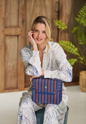 Striped blue and red handbag with textured surface and a flap closure. Model wears a white blouse with blue abstract lines, sitting on a blue stool.