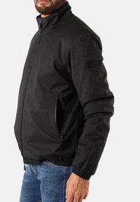Giacca in nylon nera con colletto alto, zip frontale e tasche laterali; presenta una toppa testurizzata con l'etichetta "EA7" sulla manica sinistra.
