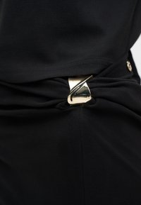 Κοντινή εικόνα μαύρου υφάσματος που συγκρατείται από έναν μεταλλικό χρυσό κρίκο σε σχήμα στυλιζαρισμένου γράμματος «Z», δημιουργώντας ένα ρυτιδωμένο εφέ.