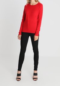 Rød langærmet sweater med rund halsudskæring, parret med sorte stramme jeans og sorte åbne hæle. Glat stof med en tætsiddende design.