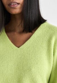 Gaišzaļš V-veida kakla džemperis, izgatavots no mīksta, strukturēta adītā auduma. Tam ir brīvs piegriezums un smalkas dūrienu detaļas ap kakla izgriezumu.