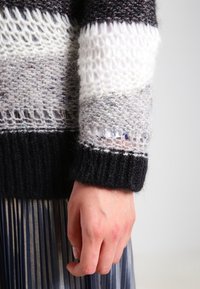 Pull en maille à rayures horizontales noires, blanches, grises et motifs texturés. Comprend des poignets côtelés et un tissage lâche pour la respirabilité.