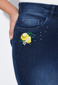 Tmavě modré džínové kalhoty s vyšitými žlutými citrony, zelenými listy a bílými akcenty, spolu s náhodně rozmístěnými kamínky.