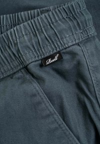 Pantalones grises con cintura elástica y una textura suave. Una pequeña etiqueta negra con texto blanco que dice "Reell" está adjunta a la cintura.