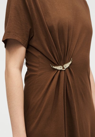 Καφέ φόρεμα με κοντά μανίκια, εφαρμοστό στη μέση με μια χρυσή καρφίτσα σε σχήμα φτερού, που δείχνει το ύφασμα να συγκεντρώνεται γύρω από το αξεσουάρ.