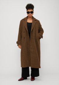 Cappotto a quadri marrone con design doppiopetto, ampie alette e due tasche laterali, indossato sopra un top nero e pantaloni neri.
