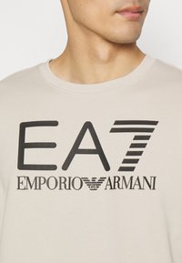 Béžová mikina s výrazným černým logem "EA7" a "Emporio Armani." Hladká textura s kulatým výstřihem a žebrovanými detaily.