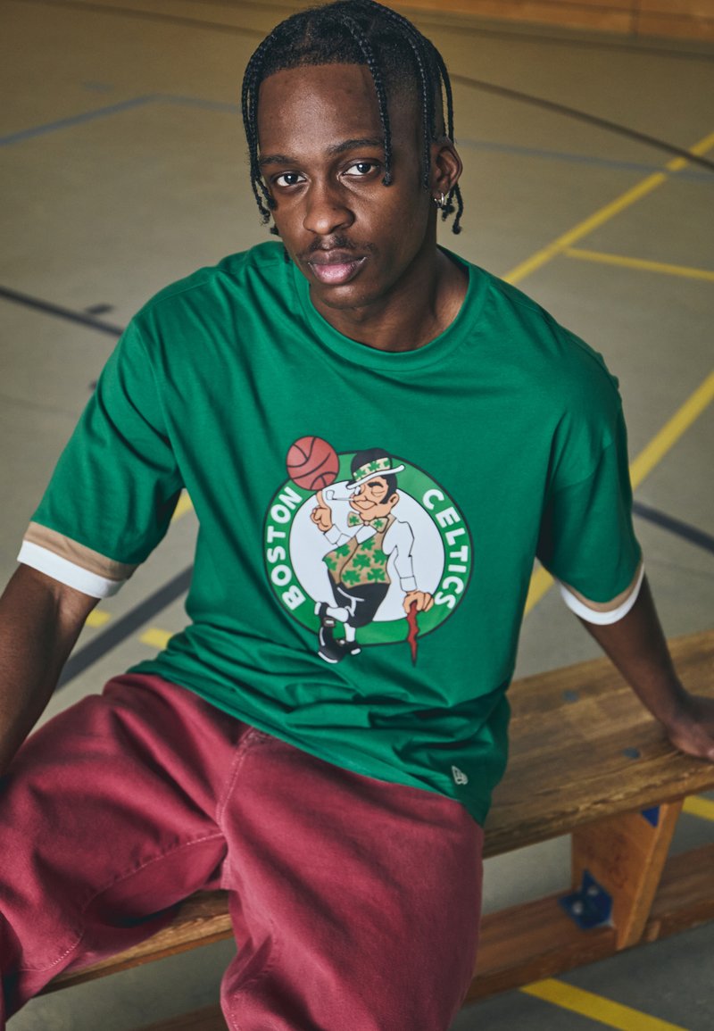 Vihreä lyhythihainen t-paita, jossa on Boston Celtics -logo, esittäen leprekaunia ja koripalloa. Päällä maroonit housut, istuen puupenkillä.
