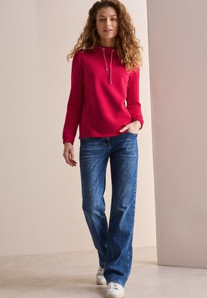 Rot pullover mit einem Kordelzug am Hals, kombiniert mit blauen Jeans. Das Model trägt weiße Sneakers und steht vor einem neutralen Hintergrund.
